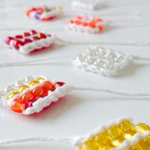 May include: A close-up of colorful beaded squares, each with a white crocheted border. The beads are arranged in a pattern of red, white, and yellow.