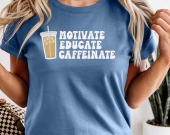Könnte beinhalten: Blaues Jeans-T-Shirt mit den weißen Worten „MOTIVATE EDUCATE CAFFEINATE“ und einer Grafik eines Eiskaffees. Das Shirt hat kurze Ärmel und einen Rundhalsausschnitt.