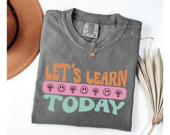 Op de afbeelding: Een grijs Comfort Colors T-shirt met de tekst "Let's Learn Today" in oranje, roze en teal. Het ontwerp bevat gloeilamp- en smiley-afbeeldingen. Een gouden ketting en een bruine hoed zijn ook zichtbaar.
