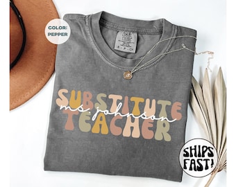 Könnte beinhalten: Ein graues Comfort Colors T-Shirt mit den Worten "SUBSTITUTE TEACHER ms. johnson" in einem Retro-Schriftzug. Eine goldene Halskette mit Anhänger befindet sich auf dem Shirt. Ein brauner Hut ist in der oberen linken Ecke sichtbar.