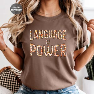 May include: Espresso-colored t-shirt with the words "LANGUAGE IS POWER" in a colorful, patterned font. The shirt is short-sleeved and has a crew neck. The overall theme is education and empowerment.