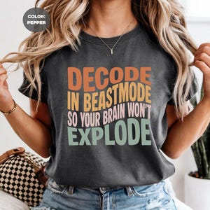 Puede incluir: Camiseta gris oscuro con el texto "DECODE IN BEASTMODE SO YOUR BRAIN WON'T EXPLODE" en naranja, amarillo, rosa y verde. La camiseta tiene cuello redondo y parece estar hecha de un material suave.