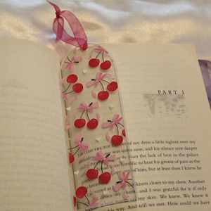 May include: A clear, rectangular bookmark featuring a repeating pattern of red cherries with pink bows. The bookmark is topped with a pink ribbon loop and is placed inside an open book, resting against a page with the printed text PART 1.