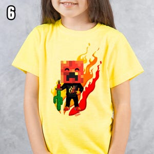 K&ouml;nnte beinhalten: Gelbes T-Shirt mit einem pixeligen Charakter mit einem feurigen Kopf, der ein Schwert und einen Kaktus h&auml;lt. Der Charakter steht vor Flammen. Die Zahl "6" ist oben links zu sehen.