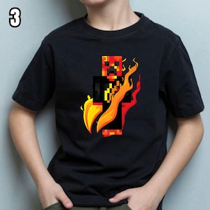 K&ouml;nnte beinhalten: Schwarzes kurz&auml;rmeliges T-Shirt mit einem pixeligen Charakterdesign in Rot, Orange und Schwarz, mit gelben Flammen. Die Zahl "3" befindet sich in der oberen linken Ecke. Das T-Shirt ist wahrscheinlich f&uuml;r Kinder.