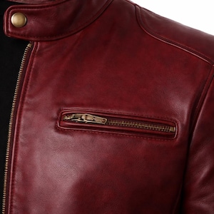 Close-up of a burgundy leather jacket with a zippered chest pocket and a gold-coloured zip. The jacket has a stand-up collar with a single gold button. The leather has a slightly distressed appearance.
