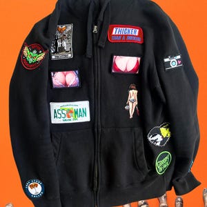 May include: Black zip-up hoodie adorned with various patches. Patches include text like "THICKER THAN A SNICKERS" and "ASS MAN", along with graphic designs. The hoodie is displayed against an orange background.