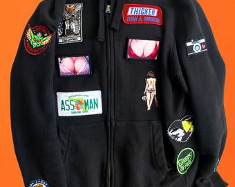 Könnte beinhalten: Schwarzer Kapuzenpullover mit Reißverschluss und verschiedenen Aufnähern. Aufnäher enthalten Texte wie "THICKER THAN A SNICKERS" und "ASS MAN" sowie Grafiken. Der Hoodie wird vor einem orangefarbenen Hintergrund präsentiert.