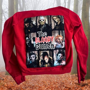 May include: Red sweatshirt with a graphic of horror film characters, including Jason Voorhees and Freddy Krueger. The phrase "The Bloody Bunch" is printed on the front. The sweatshirt is made of a soft material.