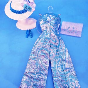 May include: A doll-sized outfit set featuring a metallic blue and silver paisley jumpsuit with braided trim at the hems, a white straw hat with blue floral and ribbon accents, a pair of blue shoes, and a set of beaded jewelry on a display card.