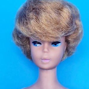 May include: A vintage doll with a blonde hairstyle and blue eyes. The doll has a neutral expression with pink lips and is set against a bright blue background. The doll's skin tone is a light beige color.