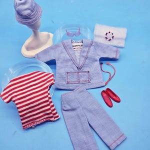 May include: A vintage Skipper doll clothing set including a light blue jacket with red stitching and a pocket, matching light blue trousers, a red and white striped shirt, a blue hat, red shoes, pink glasses, a beaded bracelet, and a white accessory.
