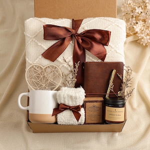 Könnte beinhalten: Eine Geschenkbox gefüllt mit einer cremefarbenen Decke mit brauner Schleife, einem braunen Lederjournal, einer Tasse, einer Kerze, einem Holzherz und einem kleinen Handtuch. Die Box wird auf einem cremefarbenen Stoff präsentiert.