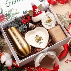 May include: A Christmas-themed gift box containing a gold tumbler, heart-shaped dish, ornament, soap, and a small Santa hat. The box is decorated with red ribbons, pinecones, and greenery, creating a festive holiday theme.