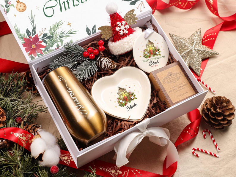 May include: A Christmas-themed gift box containing a gold tumbler, heart-shaped dish, ornament, soap, and a small Santa hat. The box is decorated with red ribbons, pinecones, and greenery, creating a festive holiday theme.