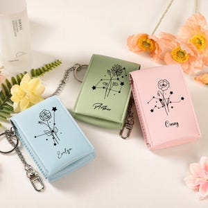 May include: Three small, rectangular keychains in pastel colors: light blue, sage green, and pink. Each keychain features a floral design with a constellation pattern and a name. The keychains have a silver chain and clasp.