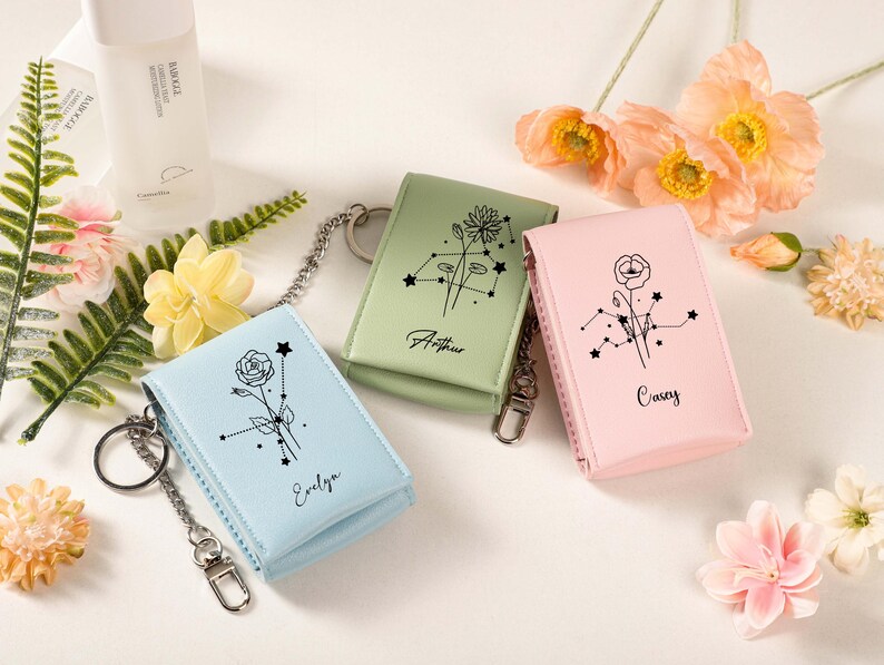 May include: Three small, rectangular keychains in pastel colors: light blue, sage green, and pink. Each keychain features a floral design with a constellation pattern and a name. The keychains have a silver chain and clasp.