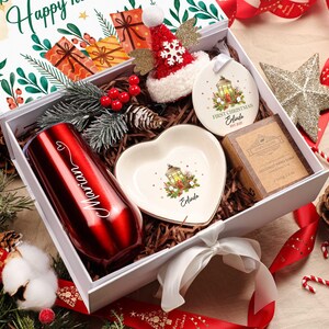 May include: A Christmas gift box containing a red tumbler, heart-shaped dish, ornament, and soap. The tumbler is engraved with a name. The box is decorated with festive elements, including a Santa hat, star, and candy canes.