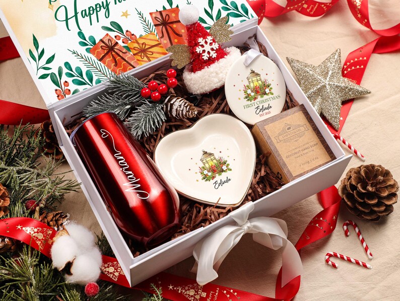 May include: A Christmas gift box containing a red tumbler, heart-shaped dish, ornament, and soap. The tumbler is engraved with a name. The box is decorated with festive elements, including a Santa hat, star, and candy canes.
