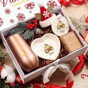 May include: A white Christmas gift box containing a rose gold tumbler, heart-shaped dish, Christmas ornament, and soap. The box is decorated with red snowflakes and a red ribbon. The box is surrounded by festive decorations.