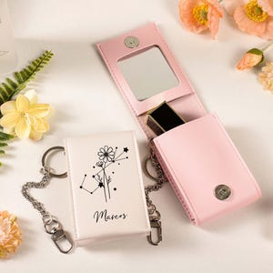 May include: A light pink leatherette lipstick case with a mirror and a silver clasp, next to a white keychain case with a floral design and the name "Marcos". The cases are on a white surface with flowers.