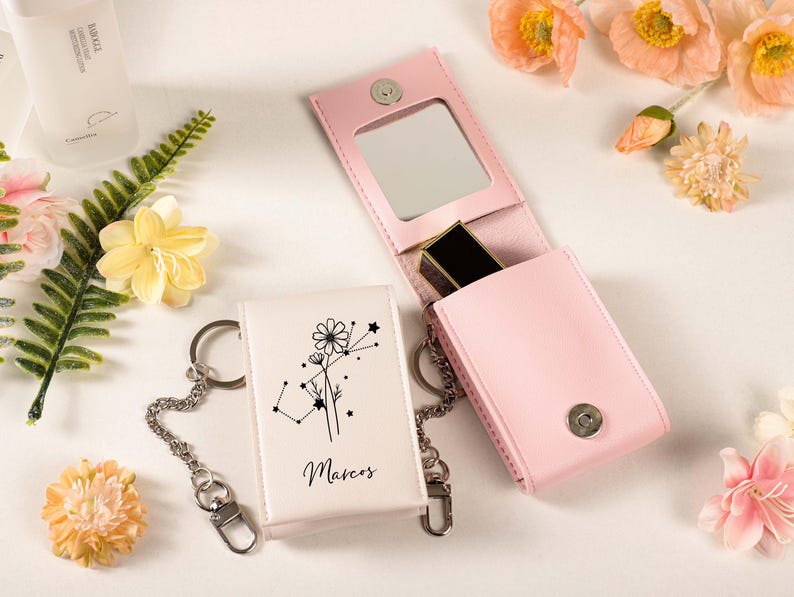 May include: A light pink leatherette lipstick case with a mirror and a silver clasp, next to a white keychain case with a floral design and the name "Marcos". The cases are on a white surface with flowers.