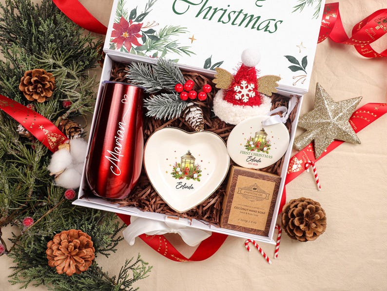 May include: A Christmas gift box containing a red tumbler, heart-shaped dish, ornament, and soap. The tumbler is engraved with "Marian". The box is decorated with greenery and a red ribbon. The ornament says "First Christmas".