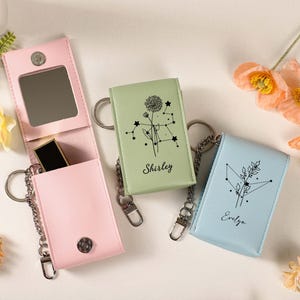 May include: A light pink leatherette keychain case with a mirror and lipstick, next to two other keychain cases in light green and blue. The green and blue cases have a constellation design and the names "Shirley" and "Evelyn" printed on them.