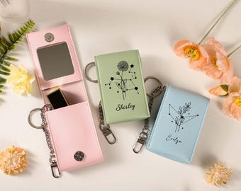 May include: A light pink leatherette keychain case with a mirror and lipstick, next to two other keychain cases in light green and blue. The green and blue cases have a constellation design and the names "Shirley" and "Evelyn" printed on them.