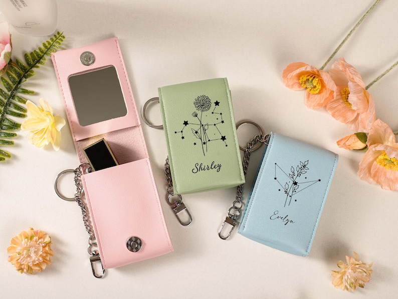 May include: A light pink leatherette keychain case with a mirror and lipstick, next to two other keychain cases in light green and blue. The green and blue cases have a constellation design and the names "Shirley" and "Evelyn" printed on them.