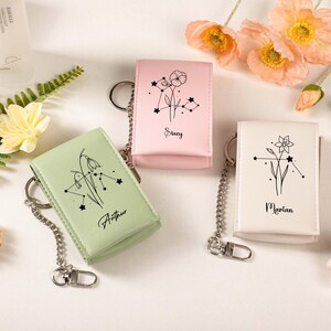 May include: Three leather keychains in pastel colors: light green, pink, and white. Each keychain features a floral design with a constellation and a name: Arthur, Stacy, and Marian. The keychains have silver chain attachments.