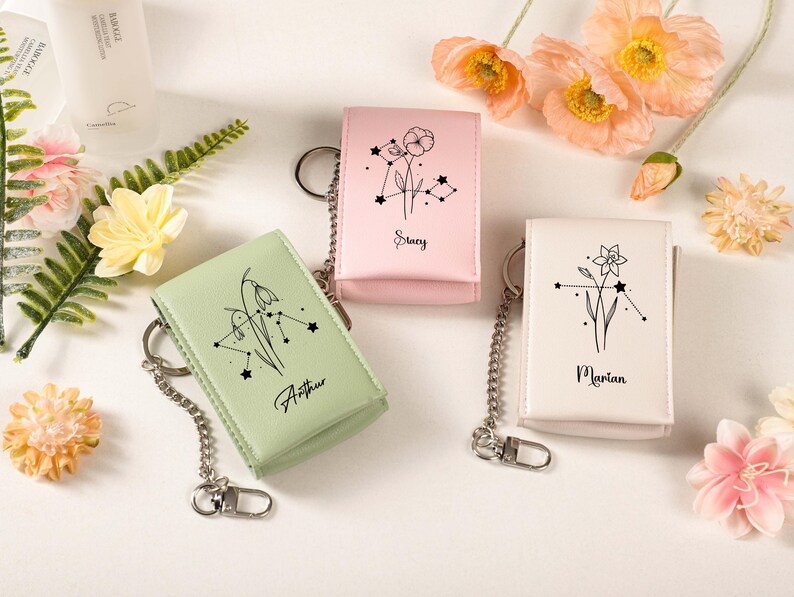 May include: Three leather keychains in pastel colors: light green, pink, and white. Each keychain features a floral design with a constellation and a name: Arthur, Stacy, and Marian. The keychains have silver chain attachments.
