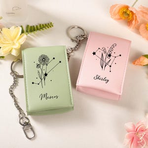 May include: Two keychains with matching cases. One is light green with a floral design and the name "Marcos". The other is pink with a floral design and the name "Shirley". Both have silver keyrings and chains.