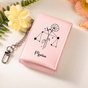 May include: A light pink wallet with a silver chain and clasp. The wallet features a black line drawing of a floral design and the Libra constellation. The name "Marian" is printed in black script below the design. The background includes flowers.