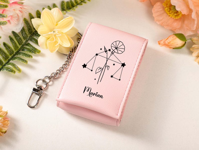 May include: A light pink wallet with a silver chain and clasp. The wallet features a black line drawing of a floral design and the Libra constellation. The name "Marian" is printed in black script below the design. The background includes flowers.