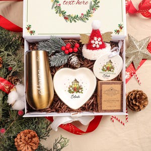 May include: A festive Christmas gift box containing a gold tumbler, heart-shaped dish, ornament, and soap. The tumbler is engraved with a name. The box is decorated with a red and white hat, pine cones, and greenery. The text "text here" is visible on the box.