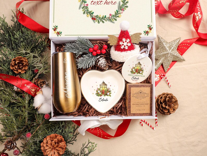 May include: A festive Christmas gift box containing a gold tumbler, heart-shaped dish, ornament, and soap. The tumbler is engraved with a name. The box is decorated with a red and white hat, pine cones, and greenery. The text "text here" is visible on the box.