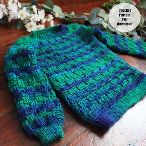 May include: A hand-crocheted sweater in shades of green and blue. The sweater features a textured stitch pattern and long sleeves. The text "Crochet Pattern PDF download" is in a white circle in the upper right corner. The sweater is displayed on a wooden surface.