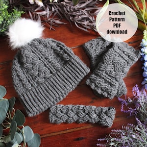 Op de afbeelding: Een set grijze haakaccessoires, waaronder een muts met een witte pompon, vingerloze handschoenen en een hoofdband. De muts en handschoenen hebben een kabelgebreid patroon. De afbeelding bevat de tekst "Crochet Pattern PDF download".