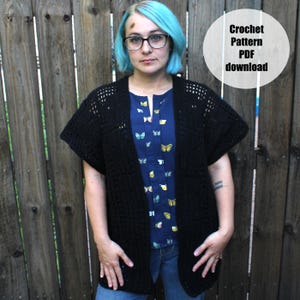 May include: A black crochet cardigan with short sleeves. The cardigan is worn over a blue blouse with a butterfly pattern. The image includes the text "Crochet Pattern PDF download" in a white circle.