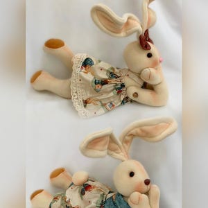 Peut inclure: Deux poupées lapins en peluche crème, l'une portant une robe et l'autre un short en jean. Les lapins ont le nez et les oreilles roses. Le texte "MOLDE COELHOS APAIXONADOS" est en haut et "por Goretti Artesanato" en bas.