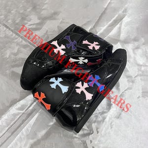 Puede incluir: Zapatillas de charol negro con detalles de cruces de colores. Las zapatillas tienen cierre de velcro y suela negra. Las cruces son en tonos rosa, azul, naranja y morado.