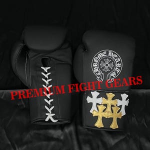 Può includere: Un paio di guantoni da boxe neri con chiusure a lacci bianchi. Un guantone mostra un logo circolare bianco Chrome Hearts, mentre l'altro presenta un gruppo di toppe a forma di croce bianche e dorate. Il testo PREMIUM FIGHT GEARS è visibile.