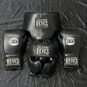 Könnte beinhalten: Ein Set schwarzer Boxausrüstung, einschließlich Handschuhen, Kopfschutz und Tiefschutz. Jedes Teil trägt das weiße "CLETO REYES" Logo. Die Artikel sind auf einer dunklen Oberfläche angeordnet und zeigen die professionelle Boxausrüstung.