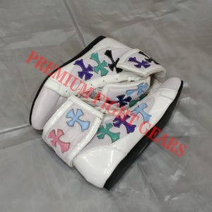 Op de afbeelding: Witte high-top boksschoenen met kleurrijke kruisontwerpen. De schoenen hebben een bovenwerk van getextureerd leer, een glanzende witte neus en een zwarte zool. De kruisontwerpen zijn in roze, blauw, groen en paars.