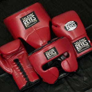 Peut inclure: Un ensemble d'équipement de boxe rouge comprenant deux gants de boxe et deux casques. Chaque article présente un écusson noir et blanc avec le logo Cleto Reyes et le texte Hecho en Mexico Professional. Les gants ont des lacets noirs.