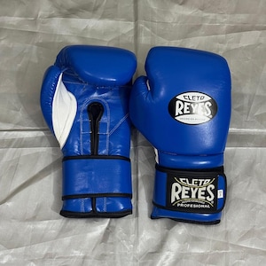 Peut inclure: Une paire de gants de boxe bleus Cleto Reyes avec des accents blancs. Les gants présentent une bordure noire, un laçage sur la paume et une sangle de poignet avec le logo Cleto Reyes et le texte 'HECHO EN MEXICO PROFESIONAL' et '14 oz'.