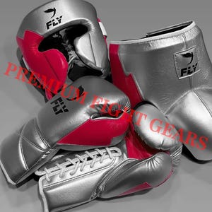 May include: A collection of silver and red boxing equipment, including a head guard, gloves, and shin guards. The gear displays the "FLY" logo and is designed for protection during training and competition. The set is made of a metallic material.