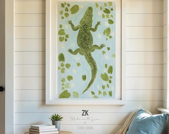 May include: A framed wall art print featuring a green alligator swimming among lily pads on a light blue background. The artwork is displayed above a small wooden side table with books and a plant, next to a wicker chair with a blue pillow.