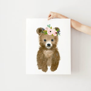 May include: A brown bear cub with a pink and green floral crown. The bear is looking at the camera with a sweet expression.